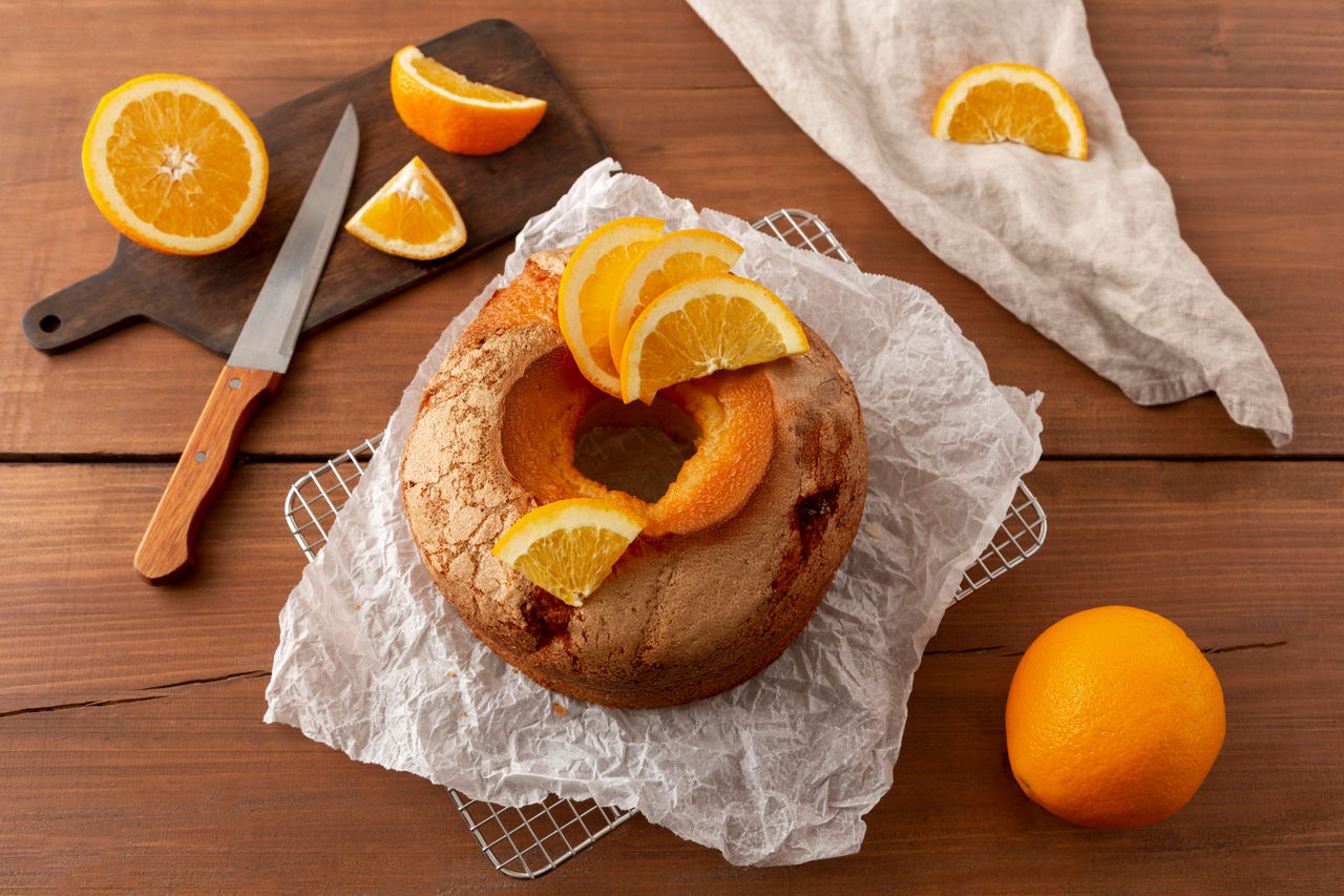 Bundt cake gegarneerd met verse sinaasappelschijfjes, omgeven door sinaasappelpartjes op een houten tafel.