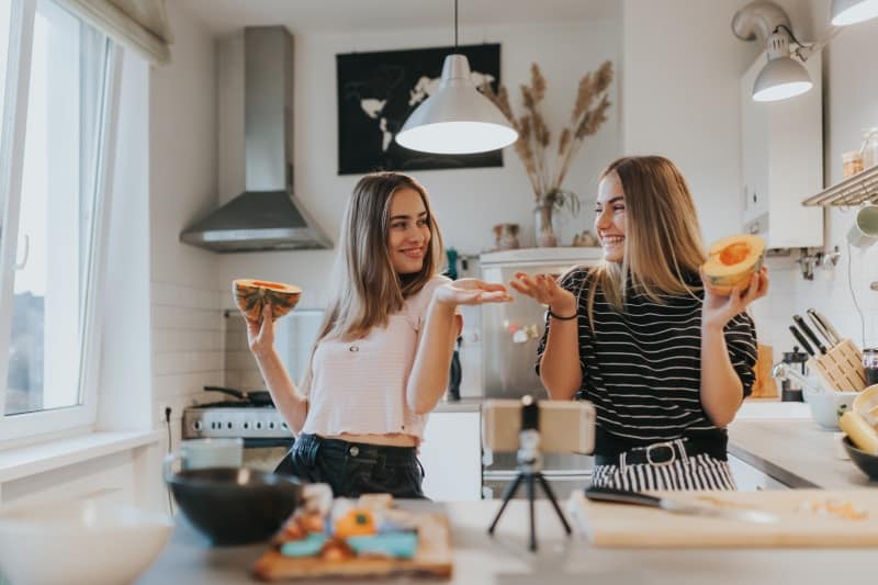 two female friends joking and eating fruit in a kitchen